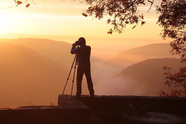 Imagen de un hombre de pie detrás de una cámara montada sobre un trípode, captando una impresionante puesta de sol con tonos vibrantes y una atmósfera serena.