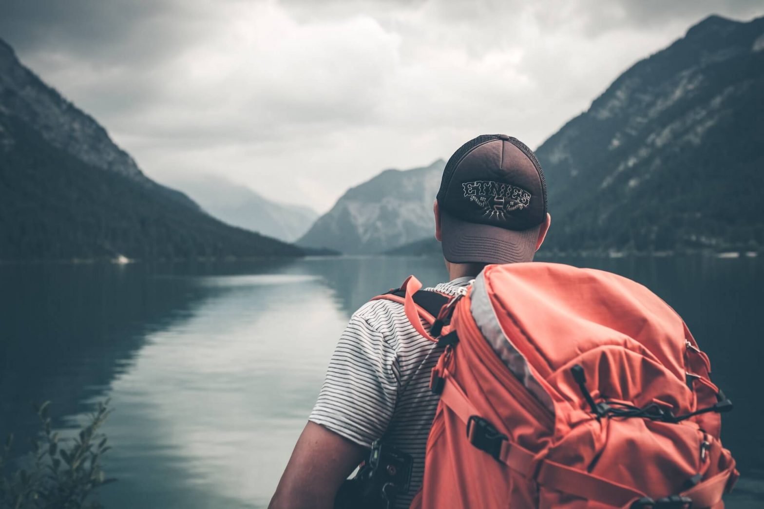 Individuo con mochila, de pie junto a un sereno lago bordeado por majestuosas montañas, capturando la belleza de la naturaleza y la exploración al aire libre.